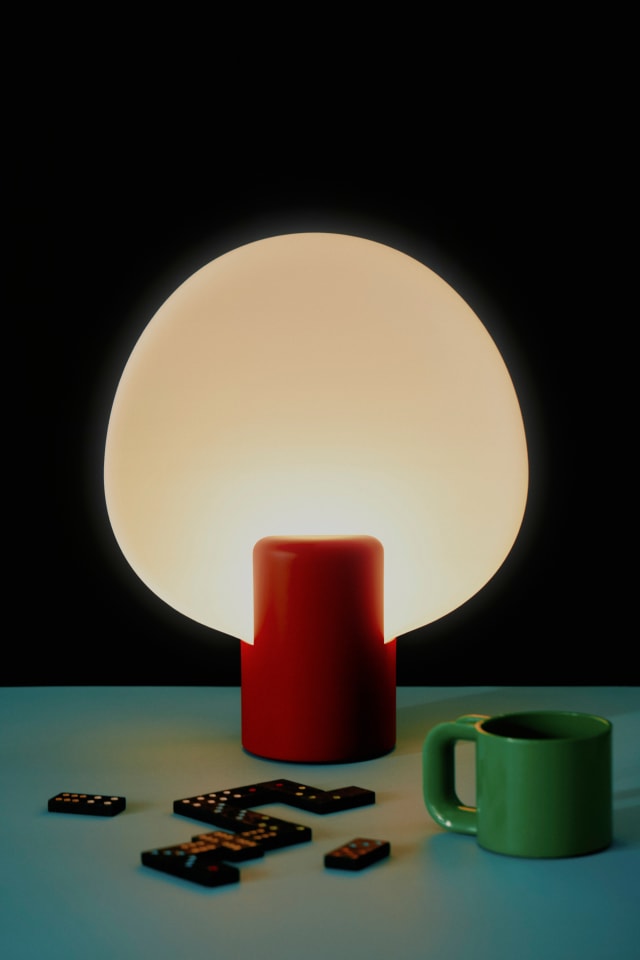A Pavone Table Light glowing beside a red mug and a green cup