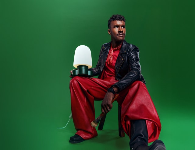 Man in a black leather jacket and red leather trousers seated against a green backdrop, holding a glowing Gantri light on his knee