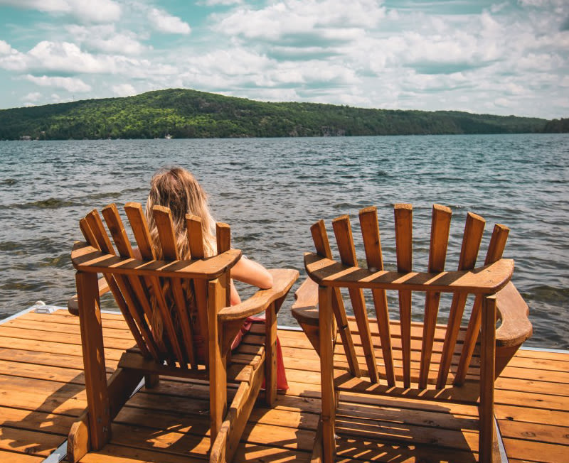 Two Adirondack chairs on a dock next to a lake with a woman sitting on the left one.