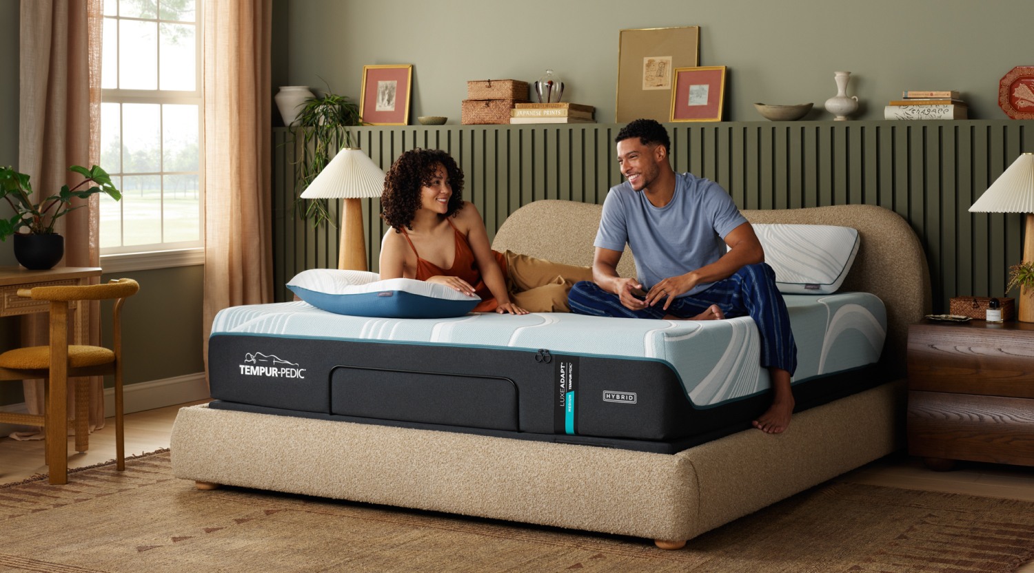 A man and woman sitting on a Tempur-Pedic queen size mattress in a bedroom.