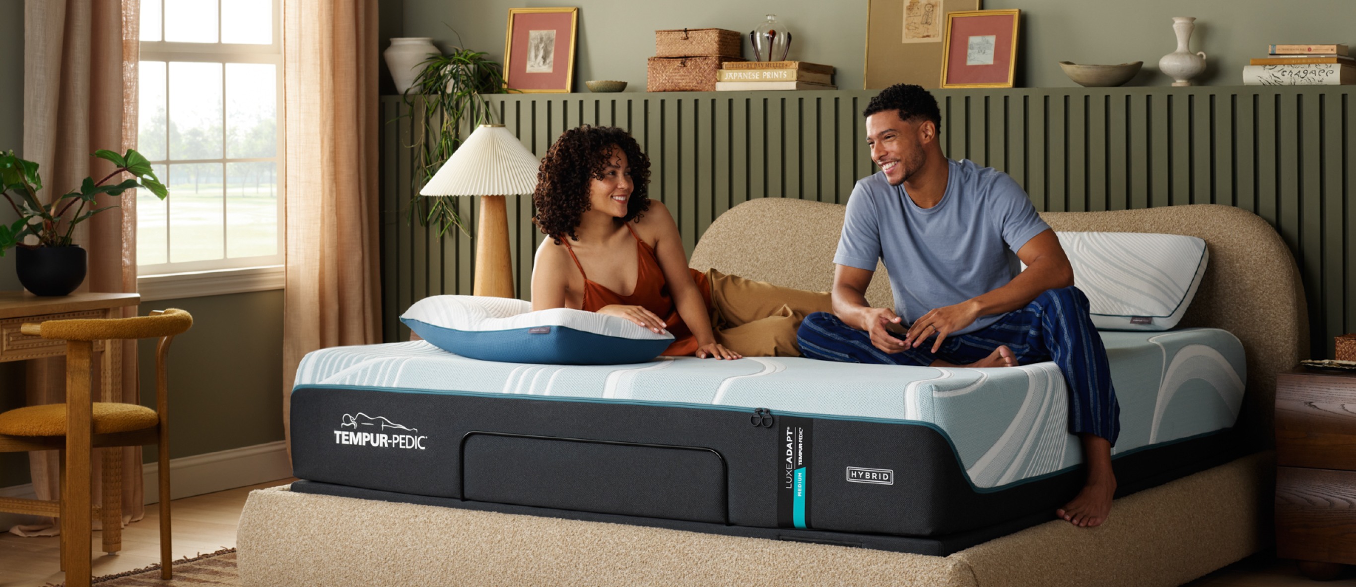 A man and woman sitting on a Tempur-Pedic queen size mattress in a bedroom.