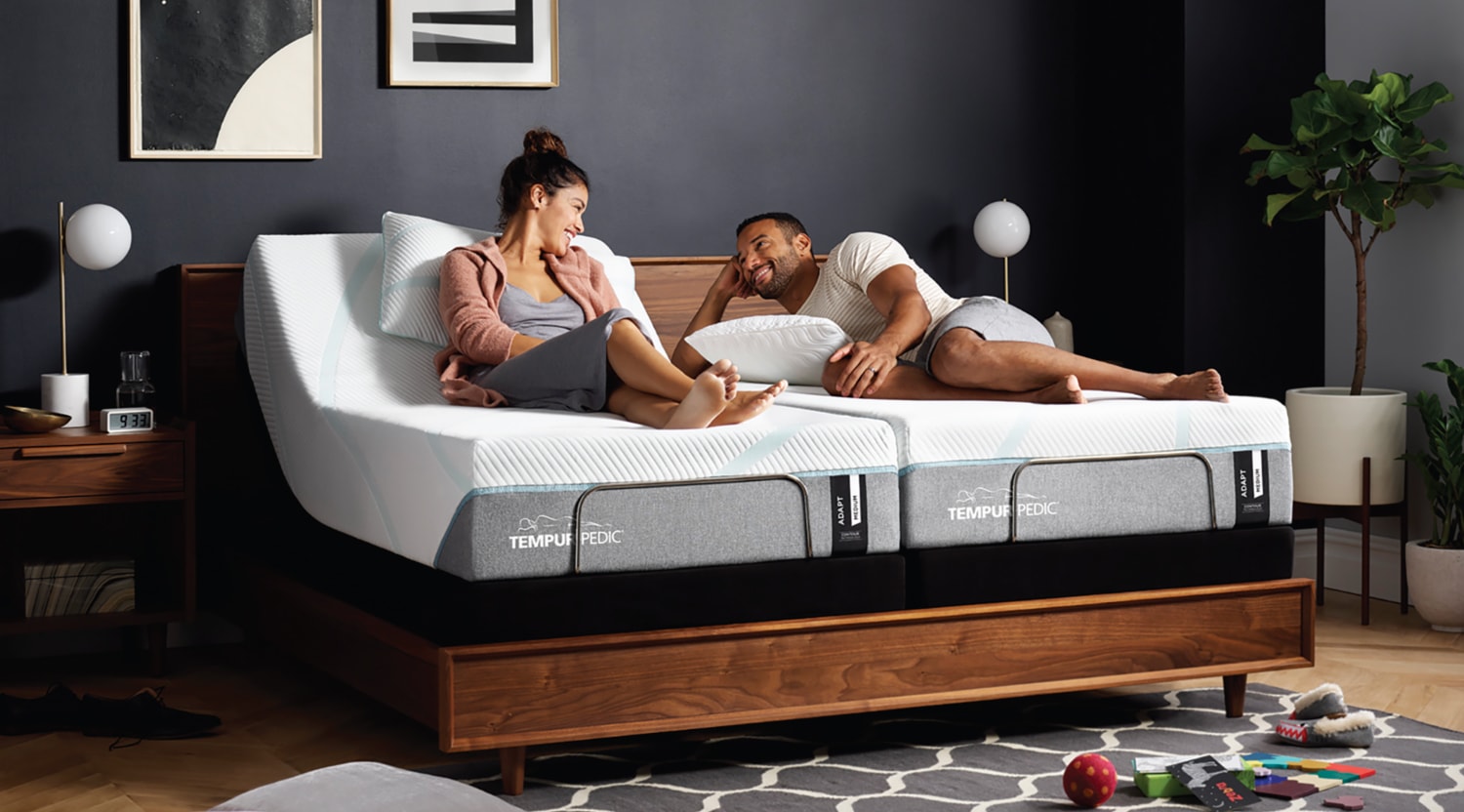 A man and woman sitting on a Tempur-Pedic queen size mattress in a bedroom.