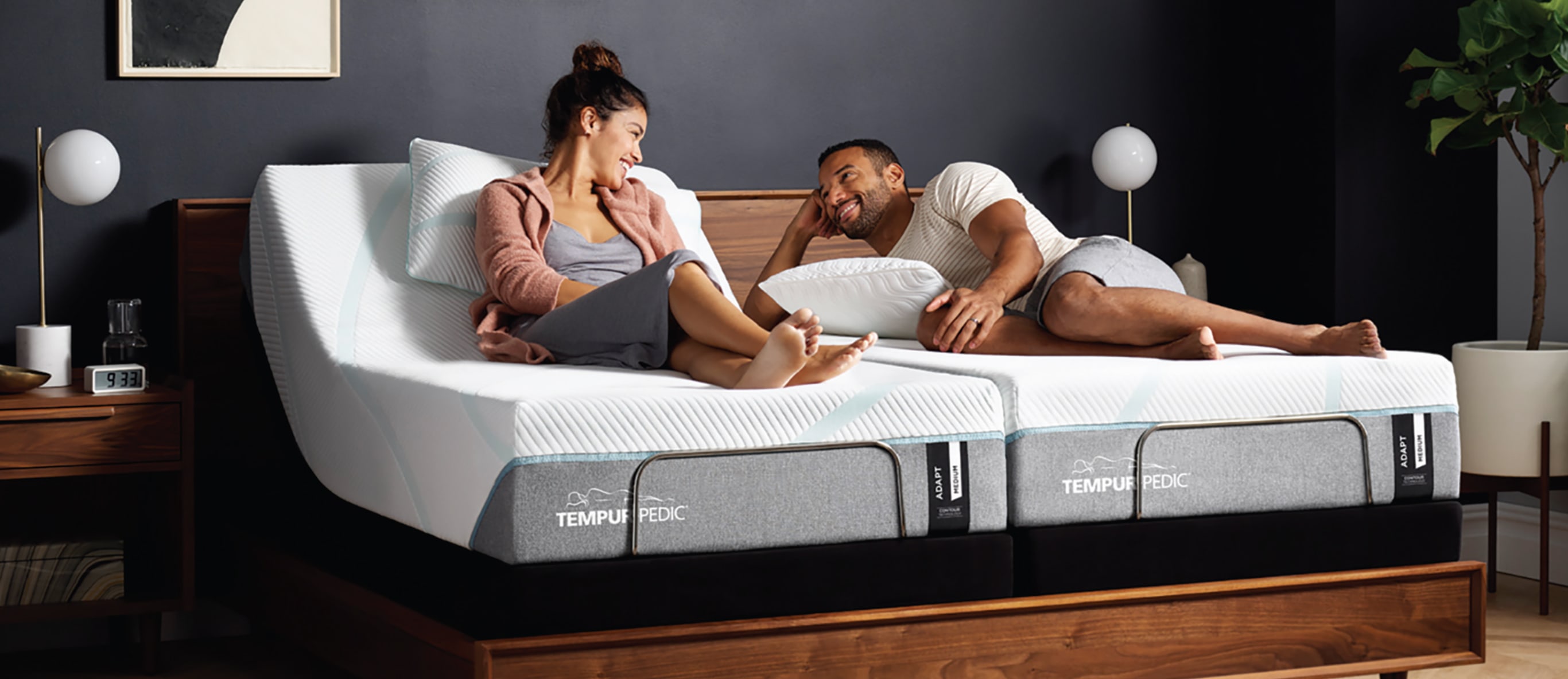 A man and woman sitting on a Tempur-Pedic queen size mattress in a bedroom.