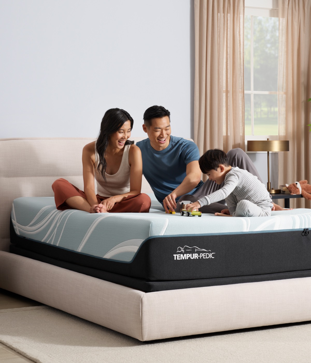 A family of three including a dad, mom, and son playing with toys atop a queen size Tempur Pedic mattress in a bedroom.