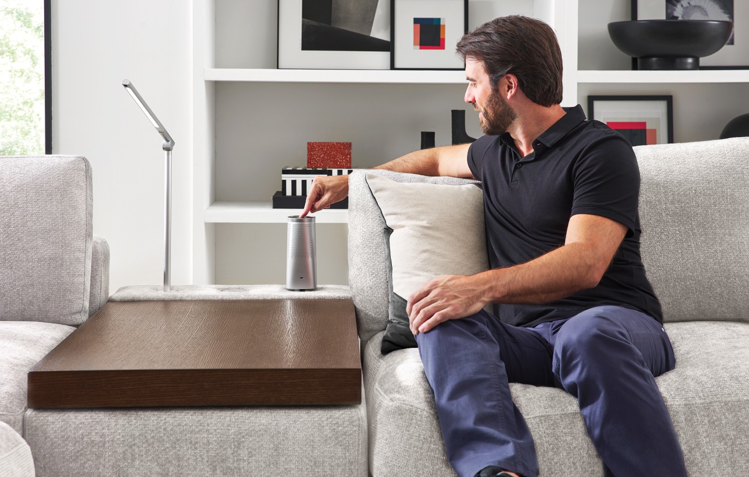 A man sits on a light gray sectional couch and manipulates the controls for audio that are built into the middle console.