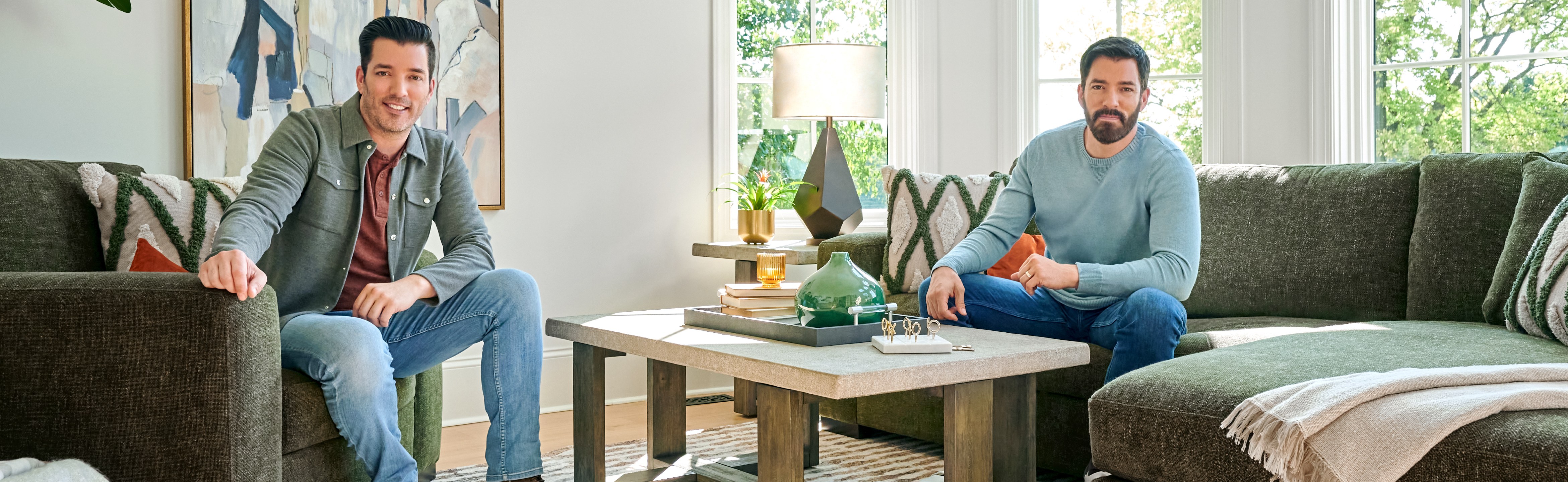 TV personalities Drew and Jonathan Scott sitting on a dark green couch and matching chair.