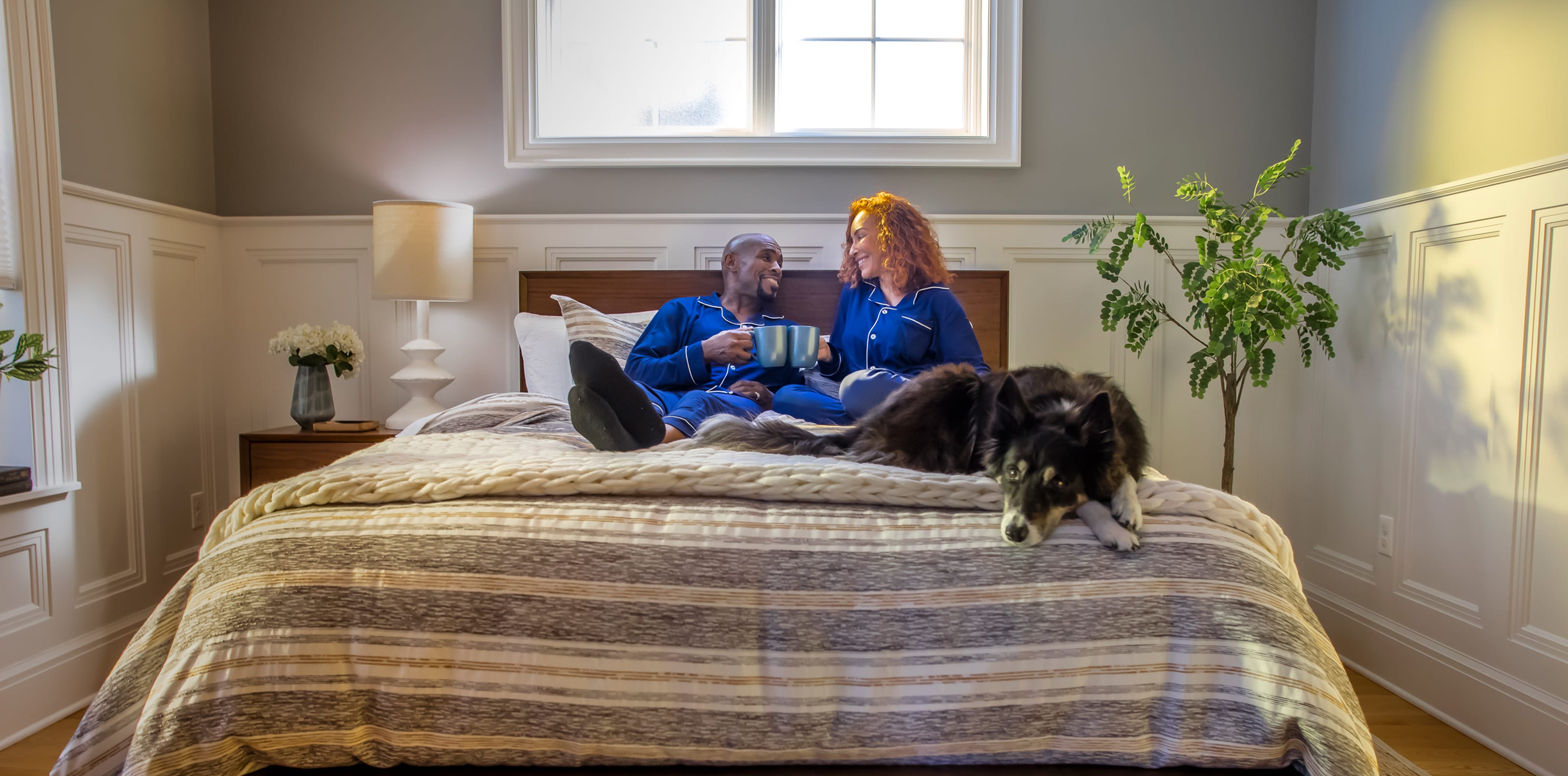 Man and woman sitting on a bed holding coffee mugs with a black and gray dog.