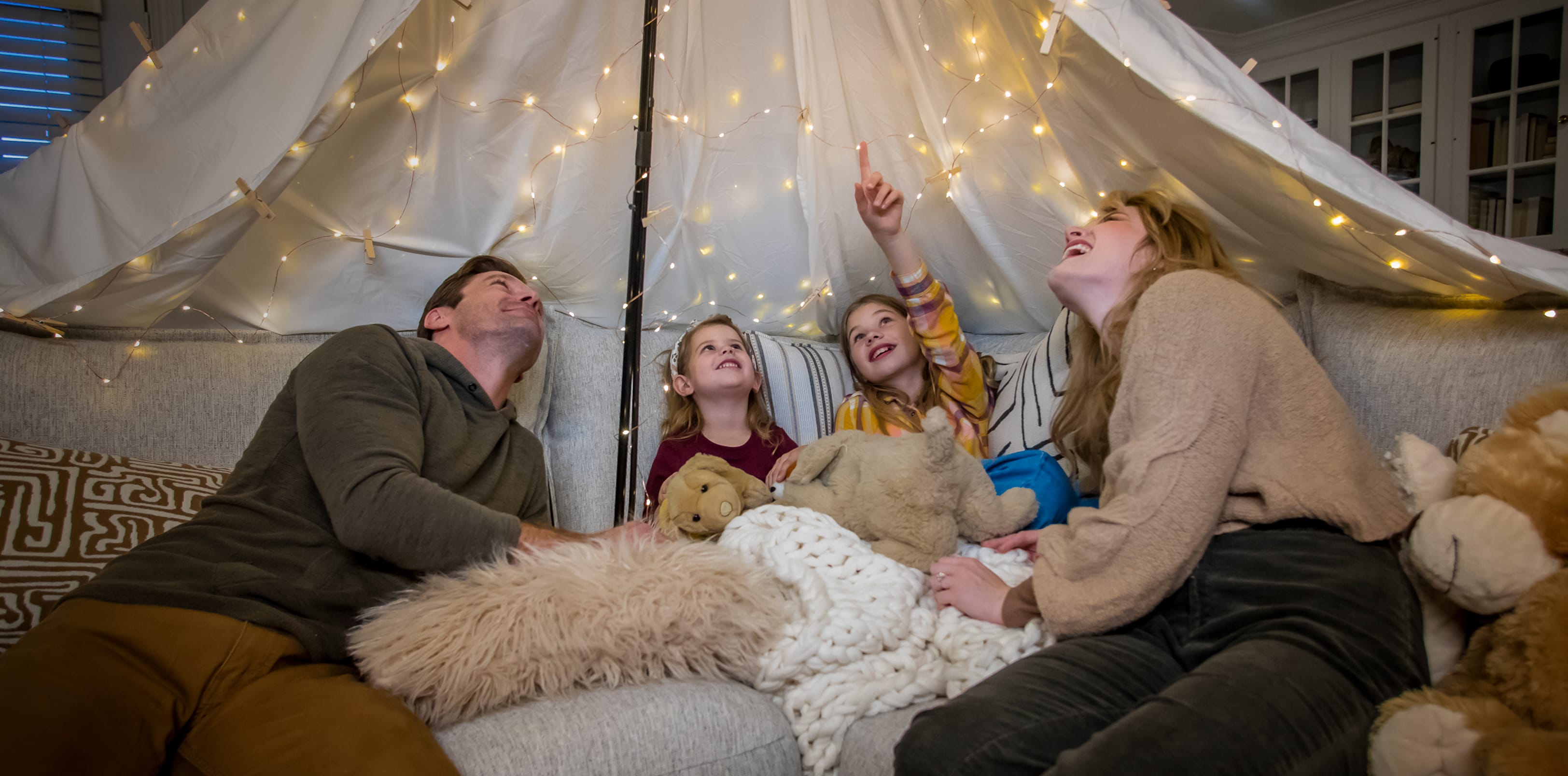 Family of 4 on a gray couch with a makeshift canopy and string lights above them.