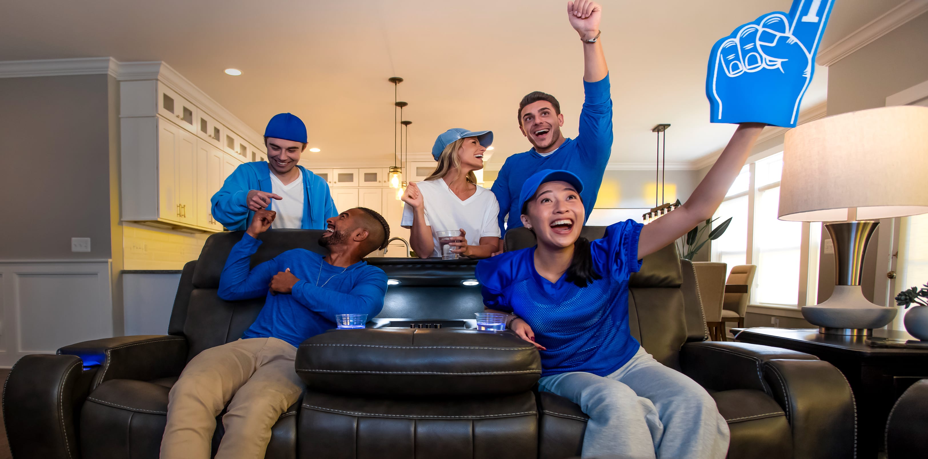 A man and woman sitting on a reclining couch wearing blue, with two men and one woman behind the couch in blue. All are cheering on a sports team.