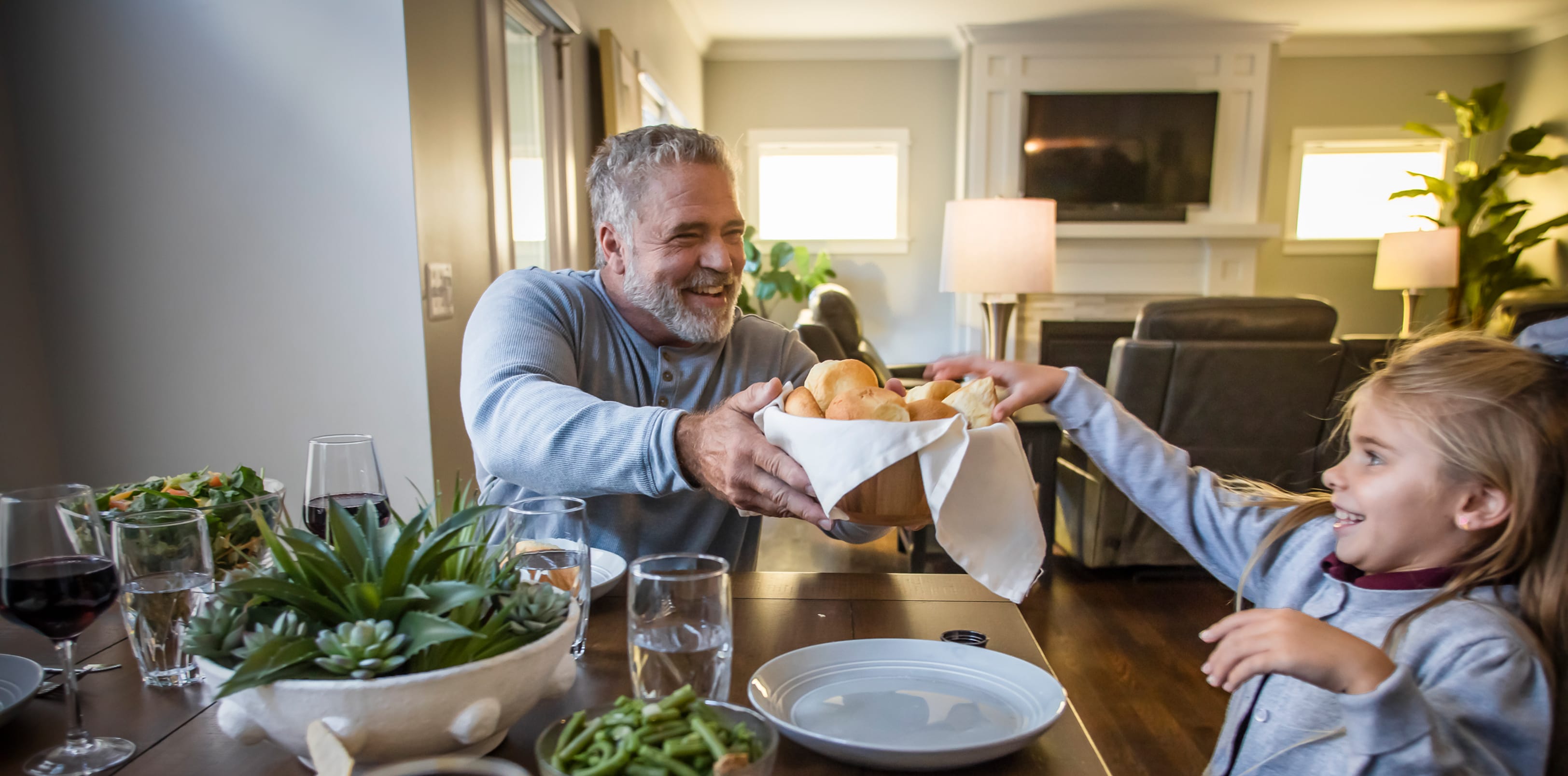 Man and child sitting at dining table passing a basket of bread rolls between them.