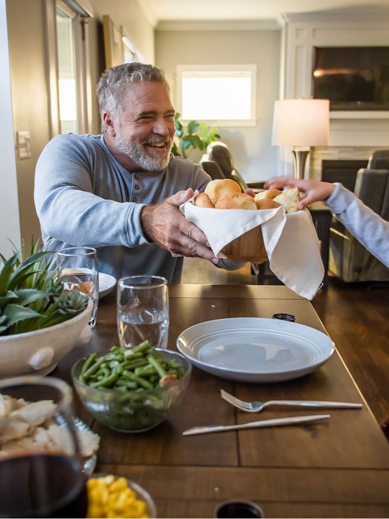 Man and child sitting at dining table passing a basket of bread rolls between them.