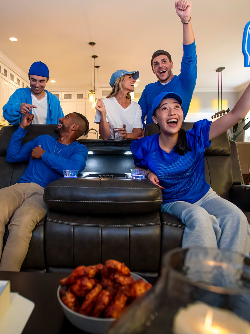 A man and woman sitting on a reclining couch wearing blue, with two men and one woman behind the couch in blue. All are cheering on a sports team.