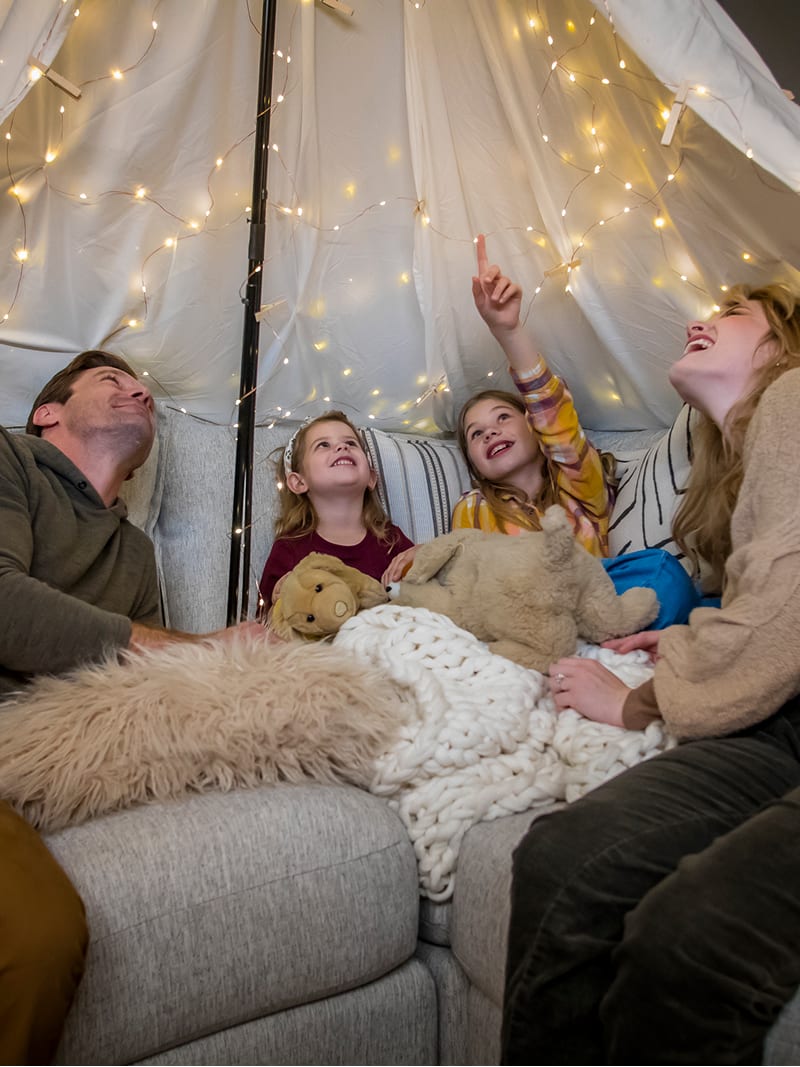 Family of 4 on a gray couch with a makeshift canopy and string lights above them.