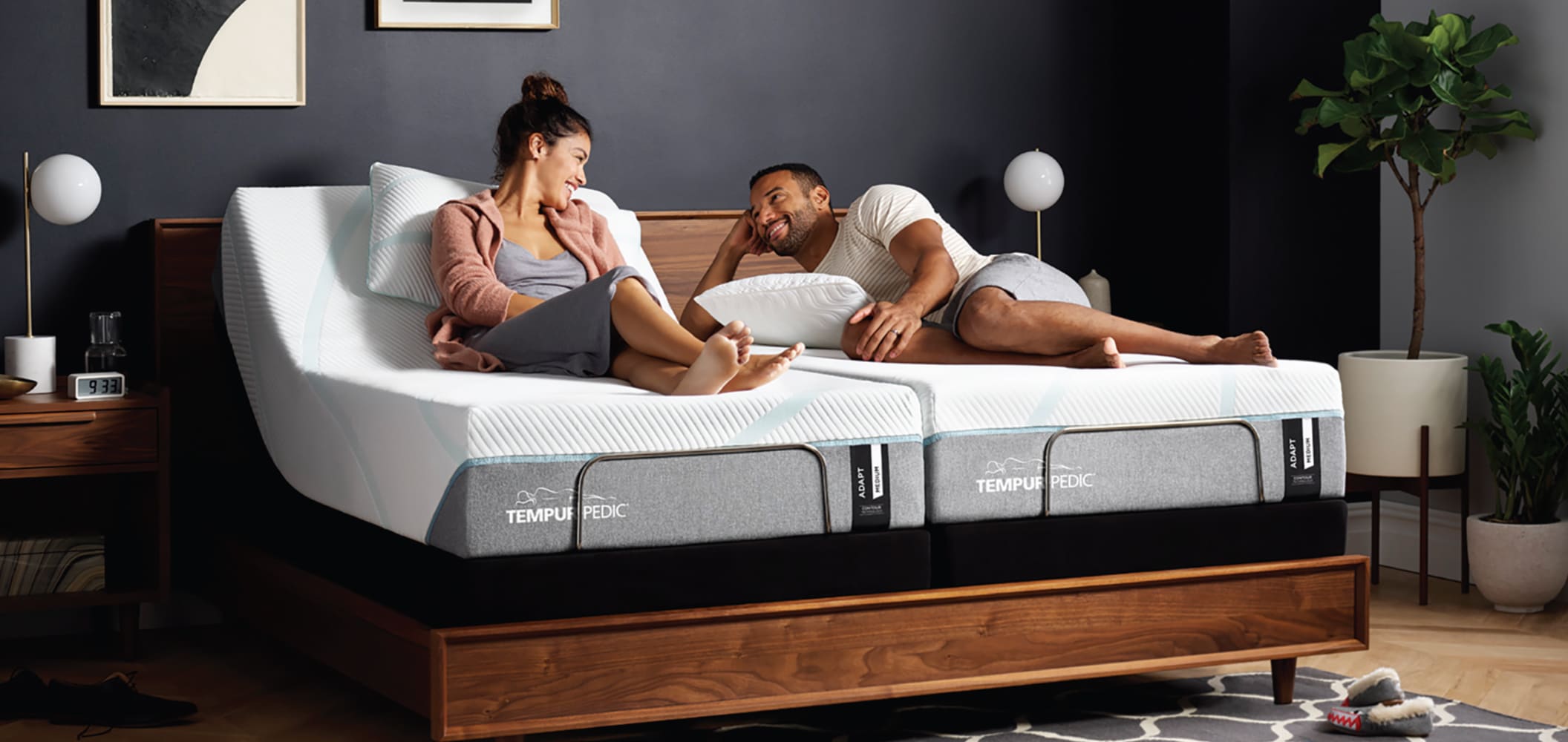 A man and woman sitting on a Tempur-Pedic queen size mattress in a bedroom.