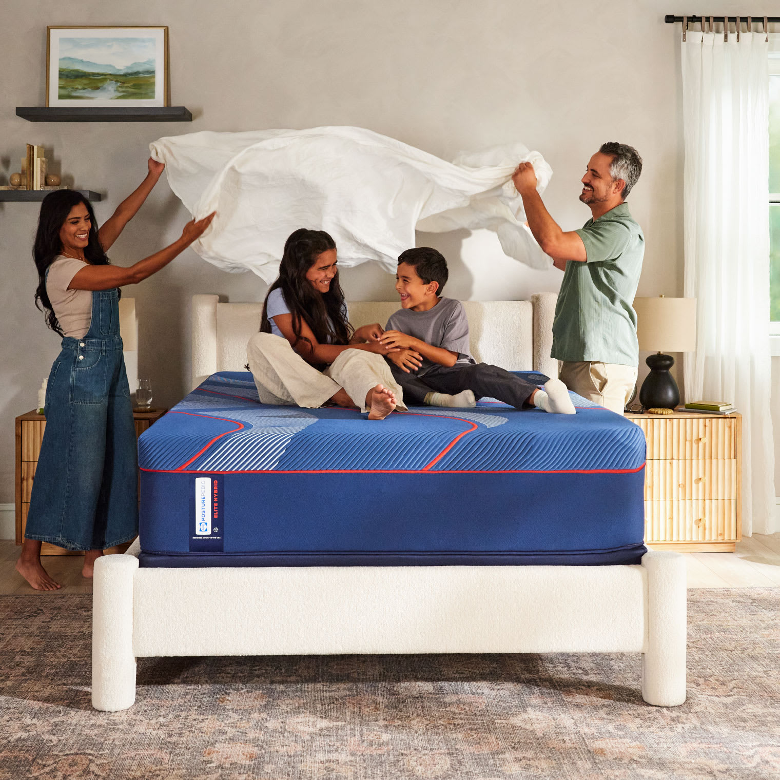 A mom and dad billowing a sheet above their son and daughter who are sitting on top of a blue queen size mattress.