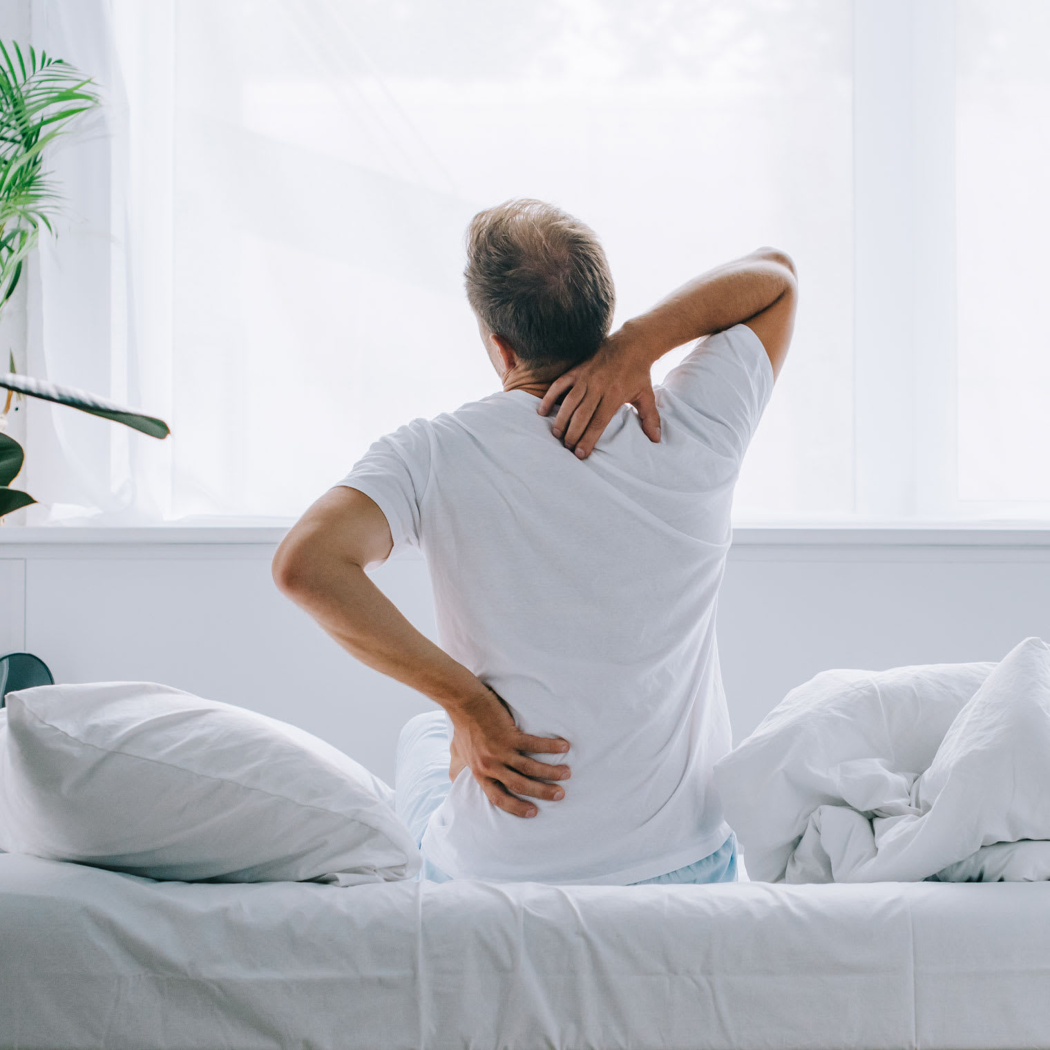 An older man with gray hair wearing a white t-shirt sitting atop a bed stretching out his back as though he is experiencing back pain.