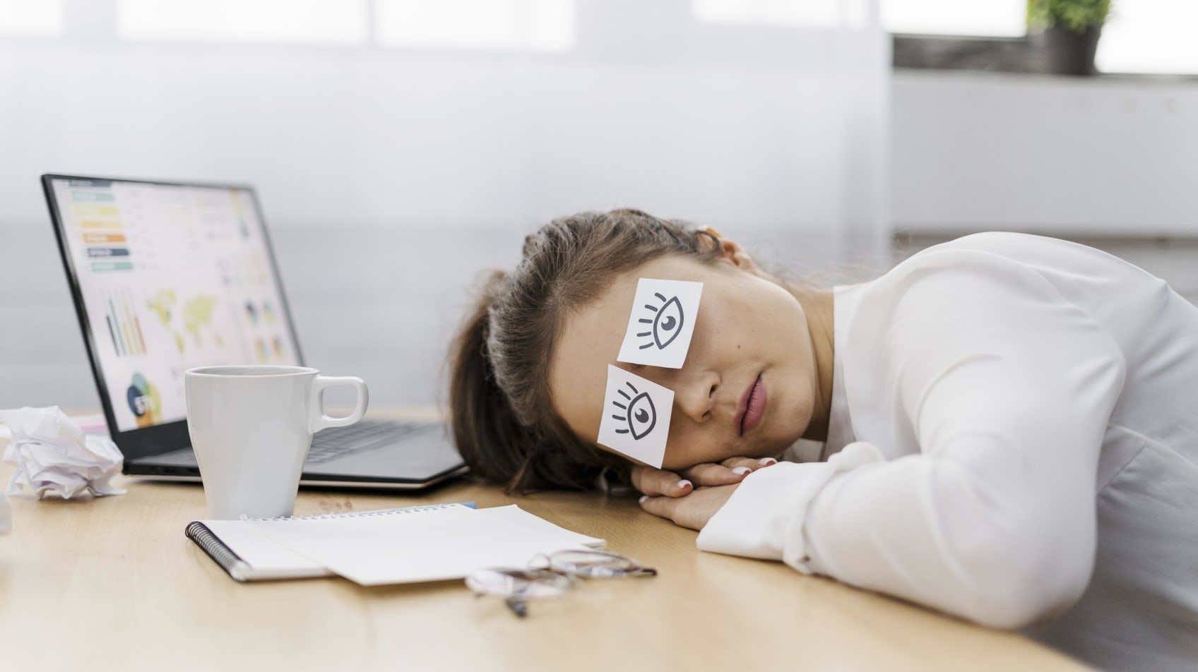 A businesswoman with her head down on her desk and pieces of paper over her eyes with open eyes drawn on them