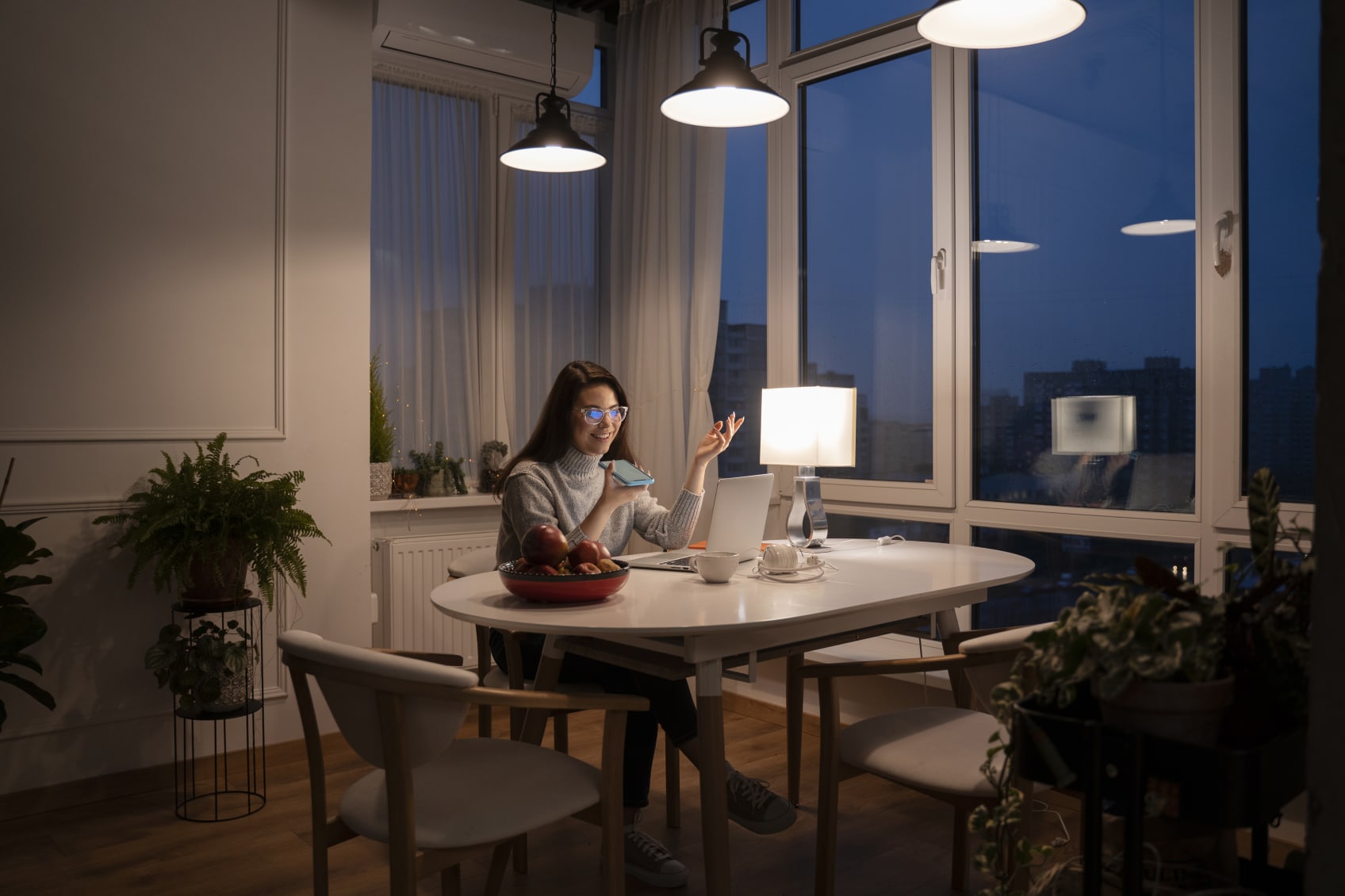 A woman talks on the phone while using a laptop at a dining table under a row of lights