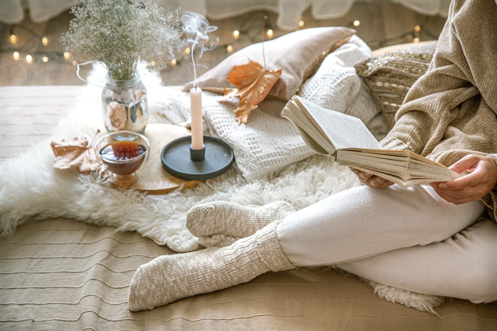 A bedroom with a lit candle, a leaf, a cup of tea, a vase with flowers in it, and a woman sitting cross-legged reading a book on a bed