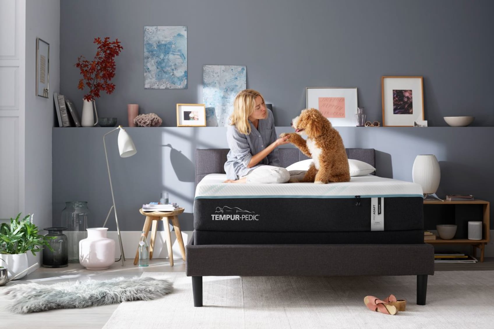 A woman shakes hands with her dog whilst sitting on a Tempur-Pedic ProAdapt Medium Hybrid mattress in a bedroom.