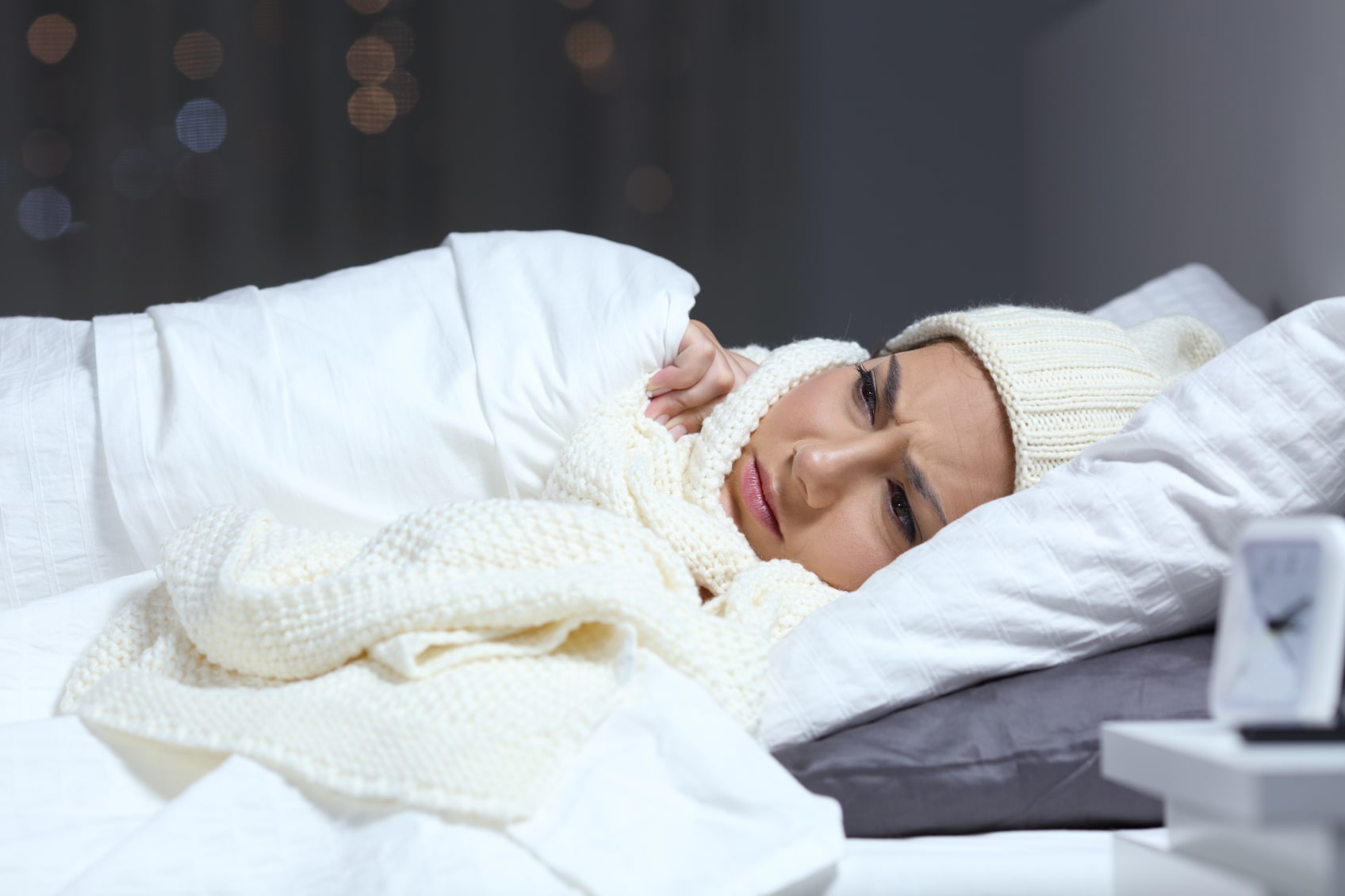 A woman in bed under the cover while also wearing a knit cap because she's too cold.