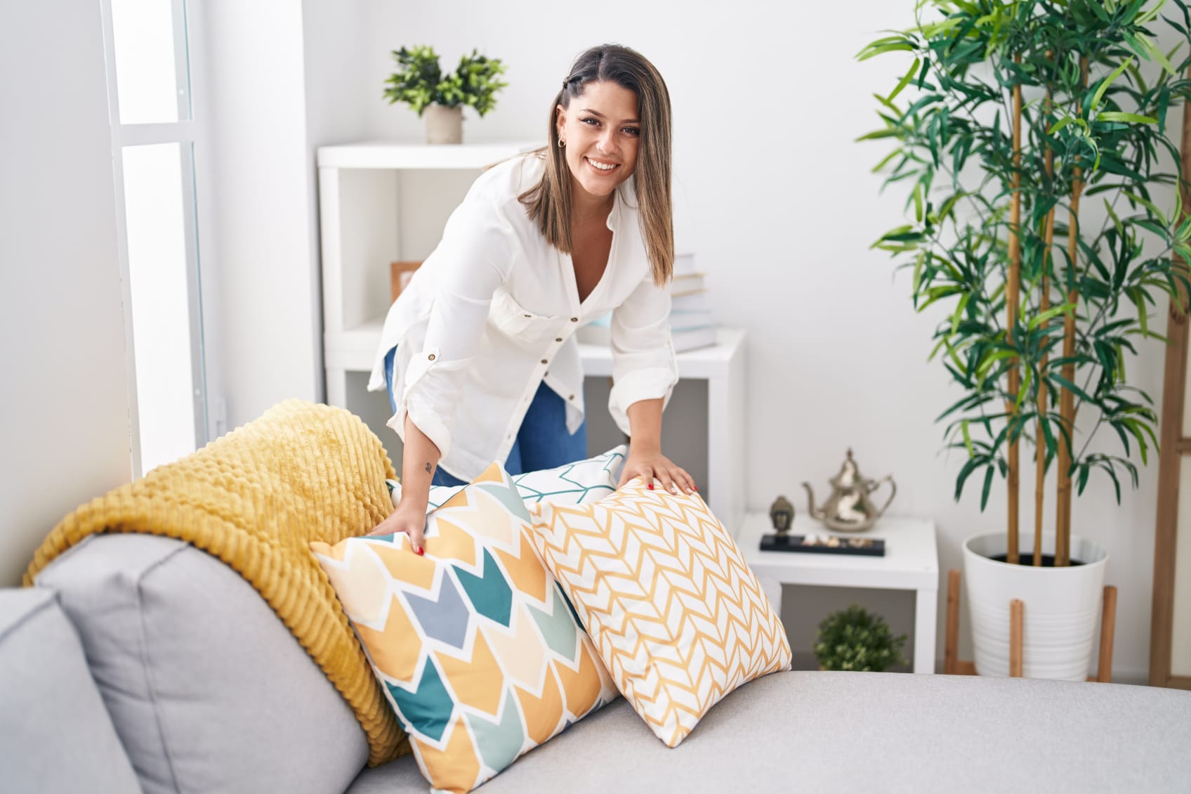 A young hispanic woman is arranging throw pillows and blankets on a couch while smiling