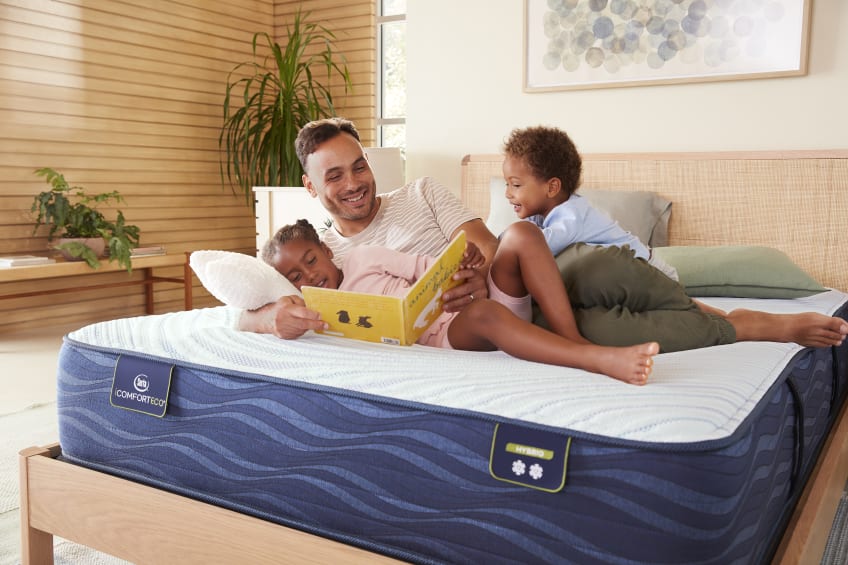 A father with his two kids lying down on a Serta mattress reading a bedtime story.