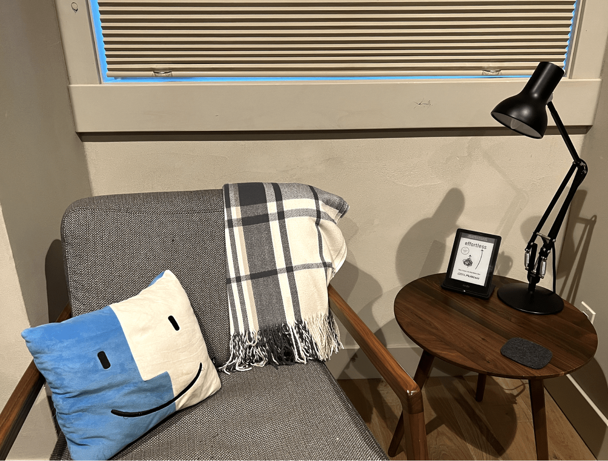 A photo of a small reading nook with a mid-century modern chair with gray cushions and small round walnut table with an adjustable lamp, a felt coaster, and a Kindle on an inclined vertical wireless charging stand.