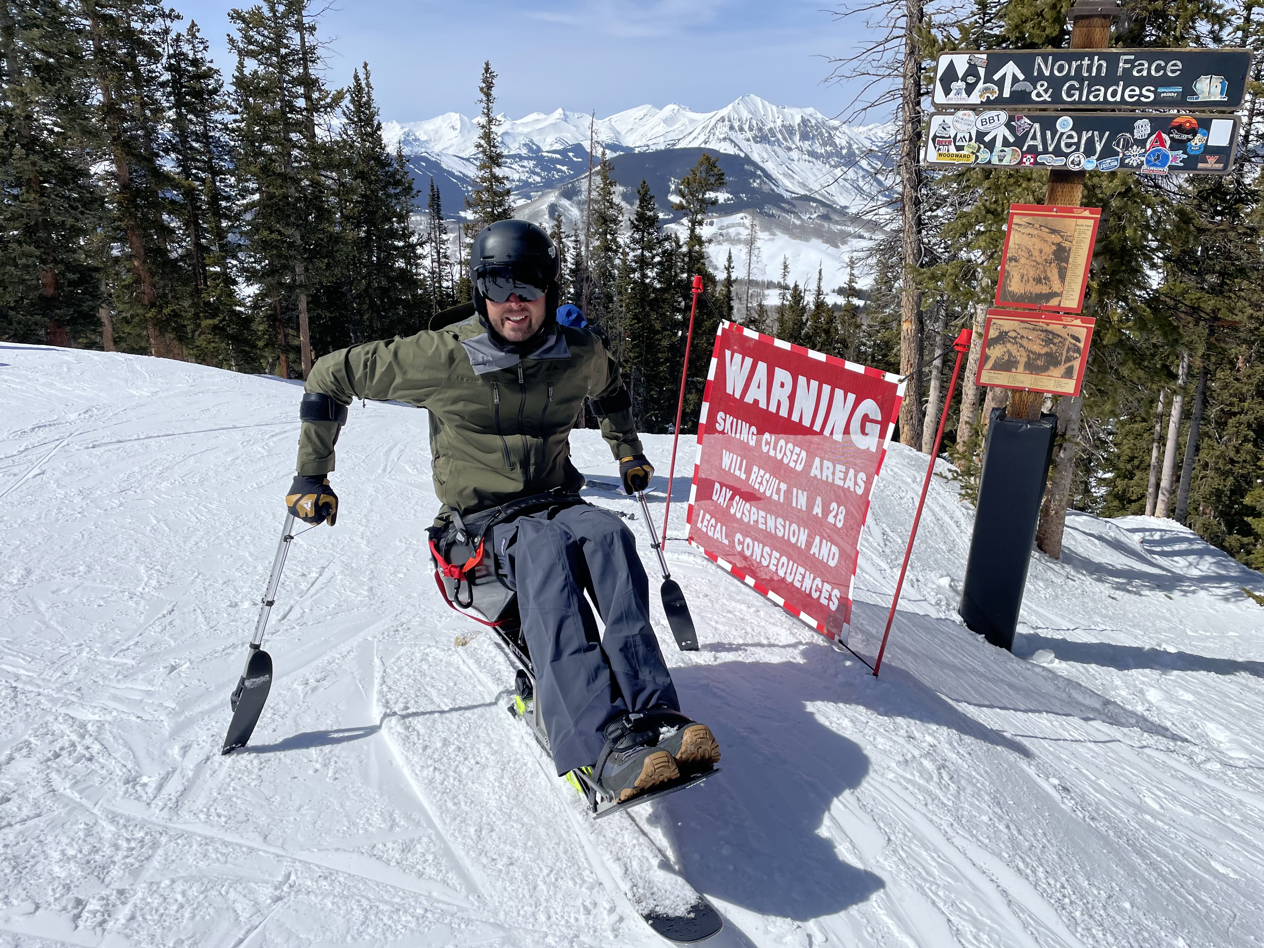A photo of Garrett in a monoski balancing with outriggers down next to a warning sign not to ski closed areas. Evergreen trees are in the near background surrounding a drop into a steep ski run. Beyond the evergreens, snow-covered mountains are in the distance.