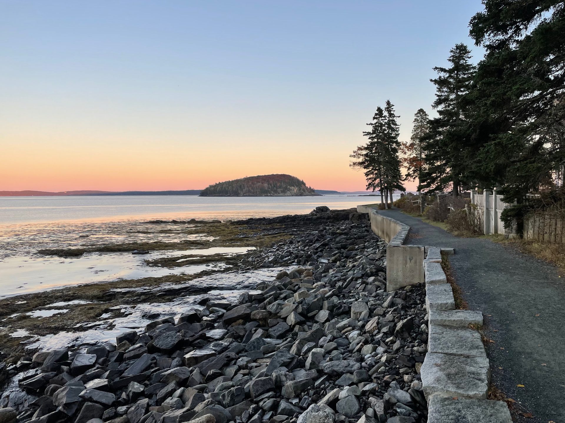 The granite-walled Bar Harbor Shore Path along the waterfront at dusk, with a wooded island visible in Frenchman Bay