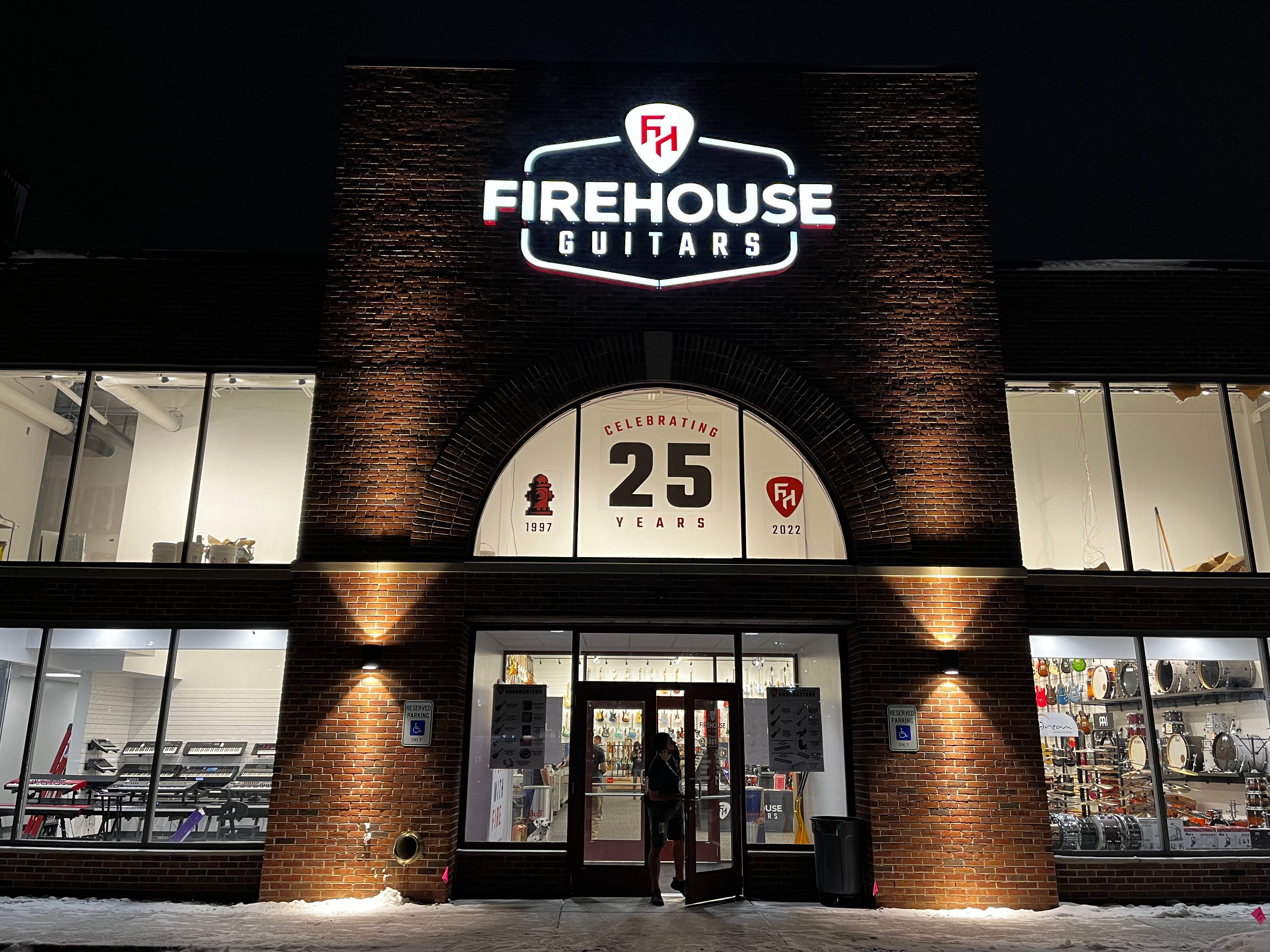Exterior of Firehouse Guitars storefront at night, featuring illuminated signage and a banner celebrating 25 years in business since 1997, with a view into the well-lit showroom displaying musical instruments.