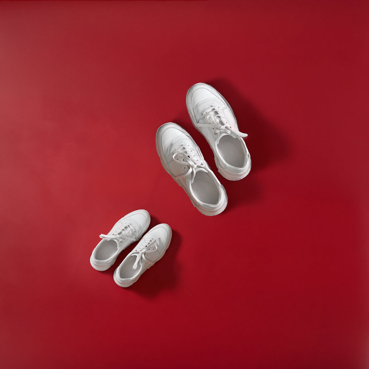 A small and a large pair of white trainers against a red background