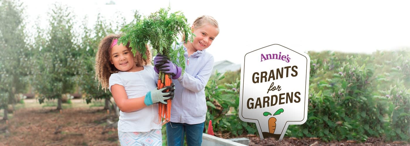 Two smiling children, holding up a big bunch of carrots, standing next to a sign that reads "Annie's Grants for Gardens"