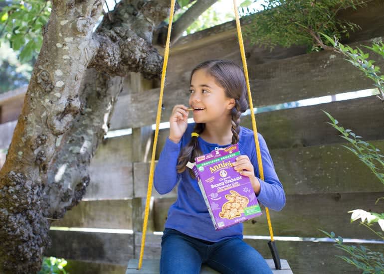 A young girl with braided hair sitting in a swing, eating Annie's Bunny Grahams from the box