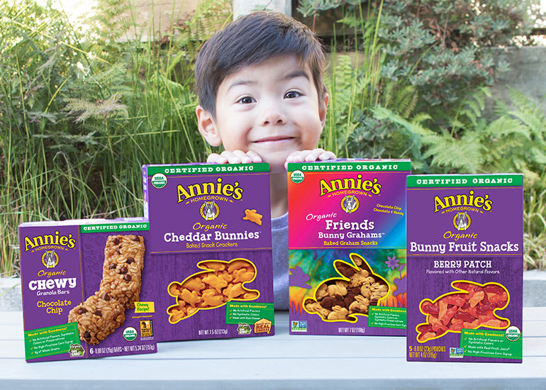 A smiling little boy peeking out from behind boxes of Annie's fruit snacks, cheddar bunnies, and granola bars
