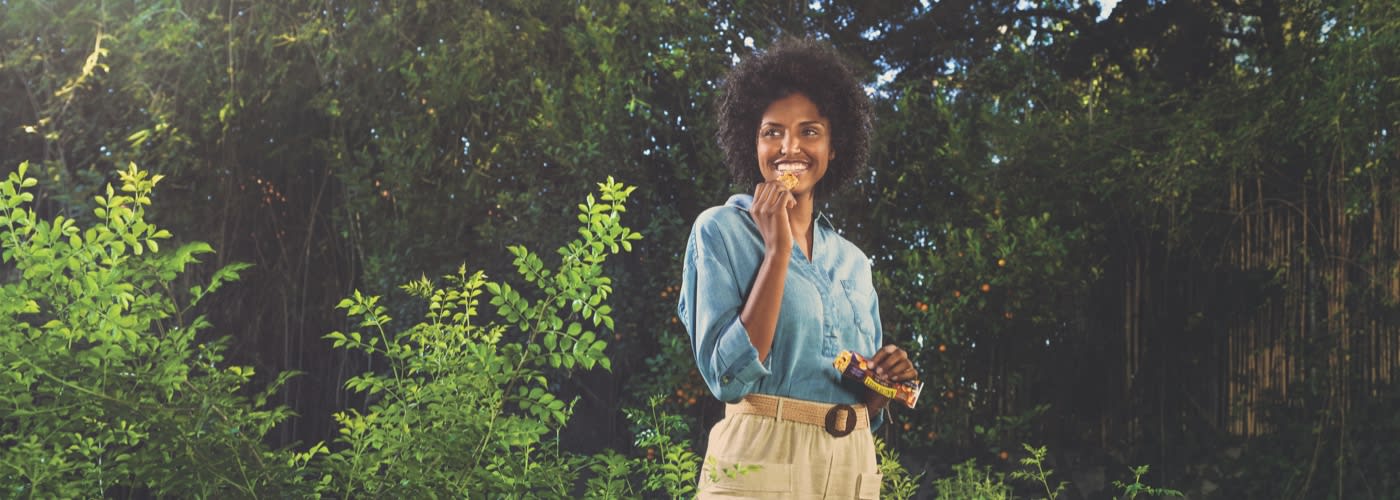 A brown with curly black hair eating Fruit and nut bars standing in front of forest