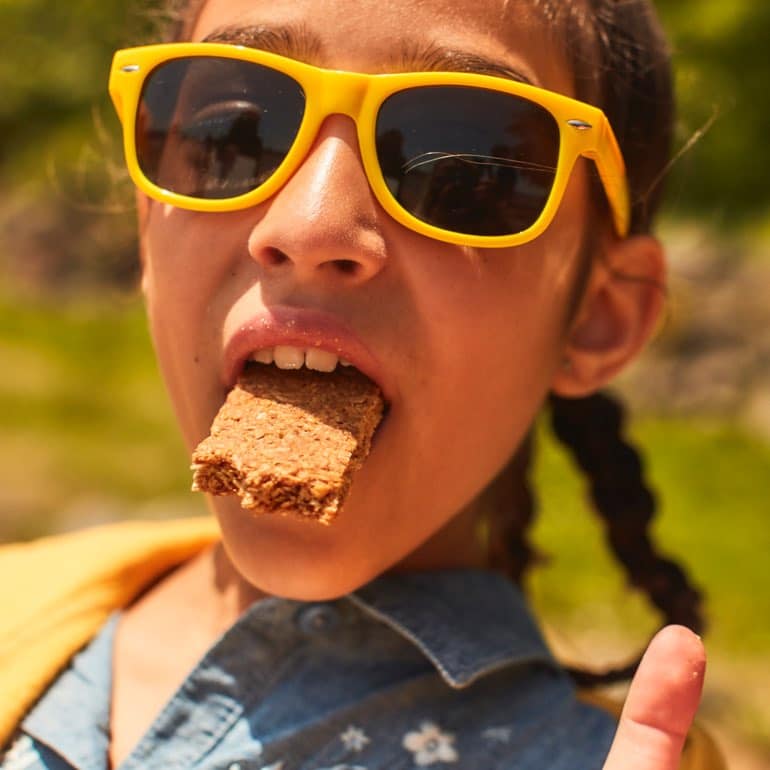 A young girl in yellow sunglasses makes a silly pose with a Nature Valley Crunchy Bar in her mouth.