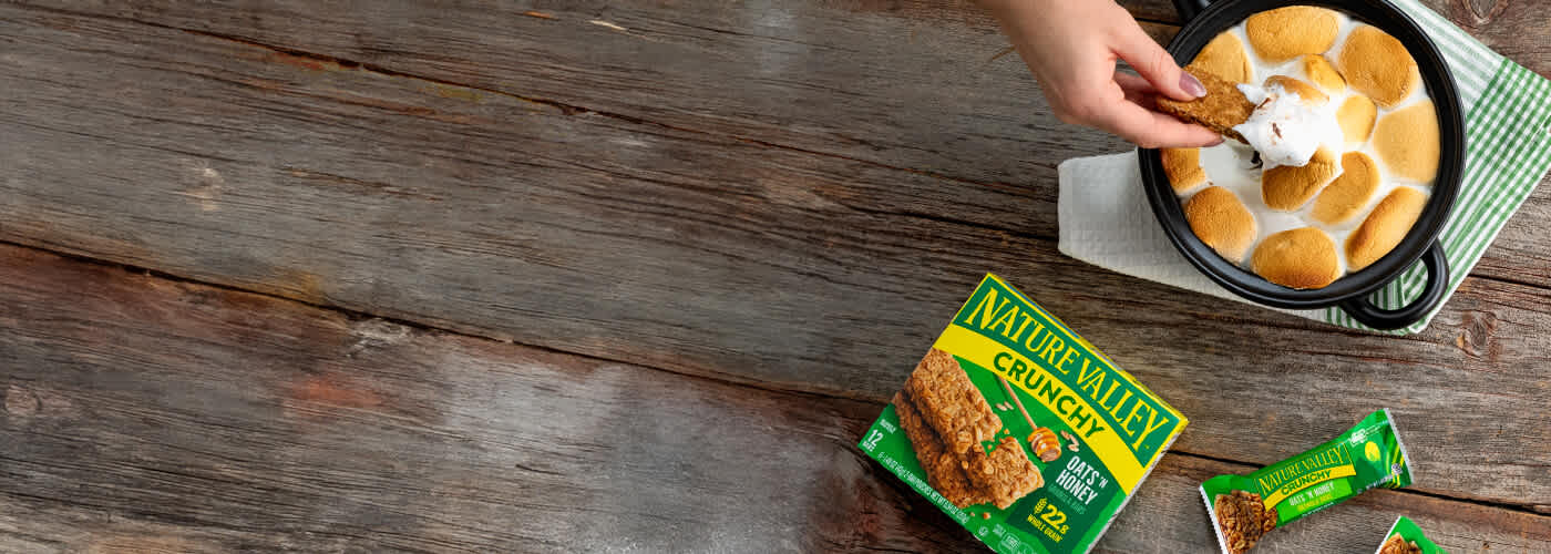 A hand dips a Nature Valley Crunchy Oats 'n Honey bar into a dish of toasted marshmallows. A green and yellow branded box and two wrapped bars are on a wooden surface.