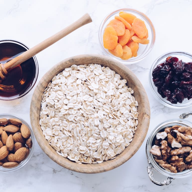 A dozen small glass bowls sitting on a marble countertop. The bowls contain ingredients such as raw almonds, pepitas, oats, walnuts, dried cranberries, coconut shavings, and dried apricots.