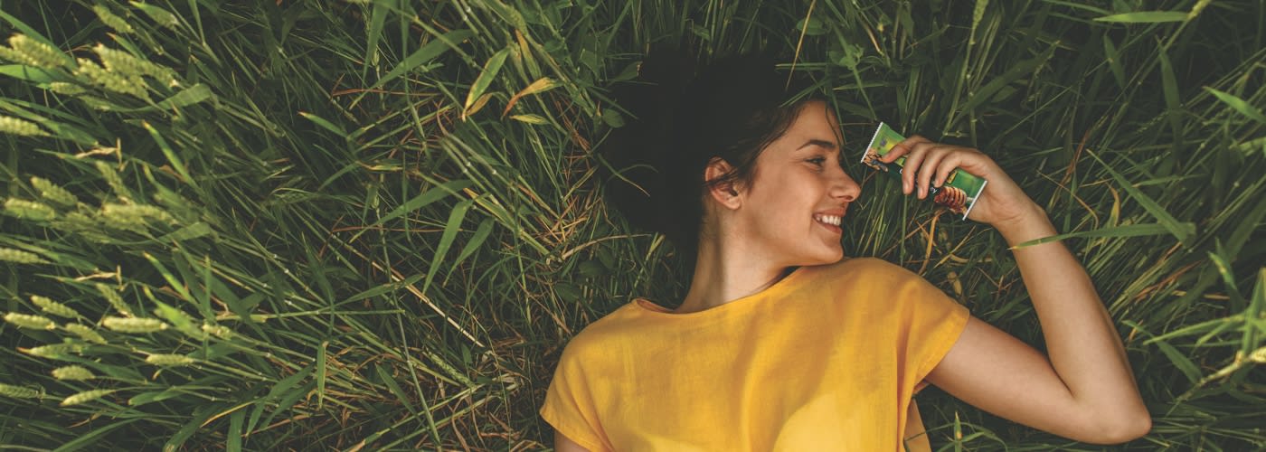 Women in Honey Color Top with Nature Valley bar in hand lying on long grass and watching the bar