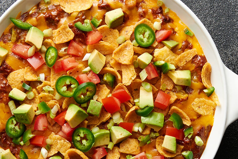 A close-up of a white skillet with Fritos Chili Bean Dip topped with chopped tomatoes and jalapenos.