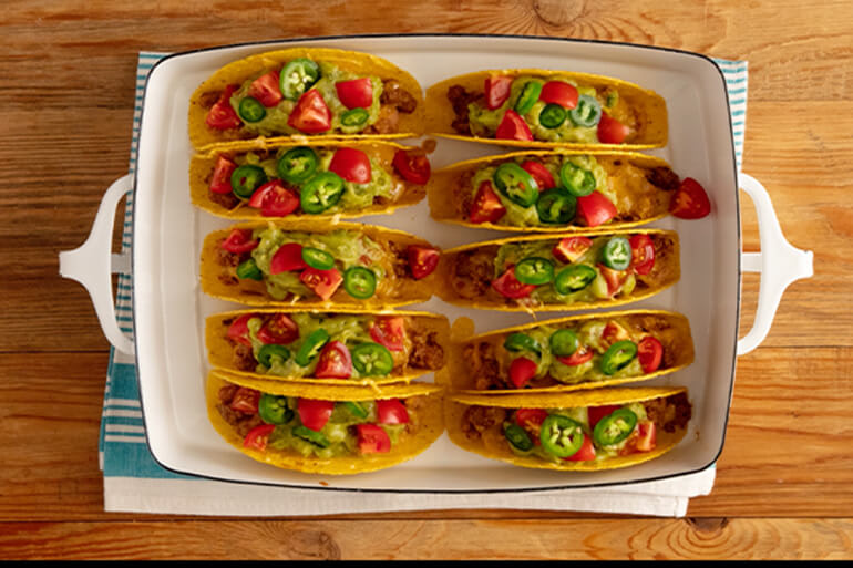 An aerial view of a white baking dish with 10 tacos lined up on a wood tabletop