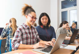 2 women sitting with their computers on a desk and looking at one laptop