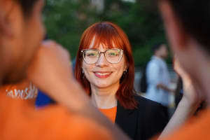 A person with red hair and glasses smiles at the camera, surrounded by people wearing orange shirts, outdoors.