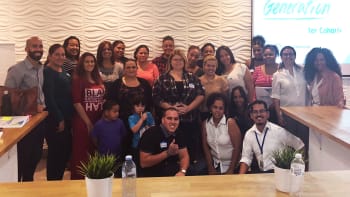 A group of about 25 people of diverse ages and backgrounds pose together indoors in front of a wavy patterned wall, with tables and water bottles in the foreground.