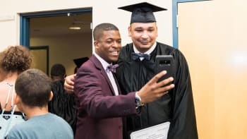 Two men pose for a selfie; one in a maroon jacket and bow tie, the other in a black graduation cap and gown holding a certificate. Other people are visible in the background.