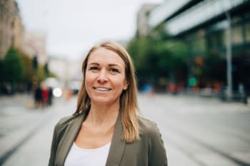 A woman with straight blonde hair, wearing a light blazer and white top, stands outdoors on a city street with blurred buildings and trees in the background.