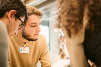 Three people sit closely together, focusing on reading a book or document. One person in the center wears a name badge and a beige sweater. The setting appears to be indoors.
