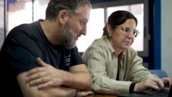 Two people sitting at a table looking at a laptop, with one person typing and the other observing closely.