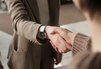 Two people shaking hands, one wearing a brown suit and a digital wristwatch, and the other in a knitted sweater, in an indoor setting.